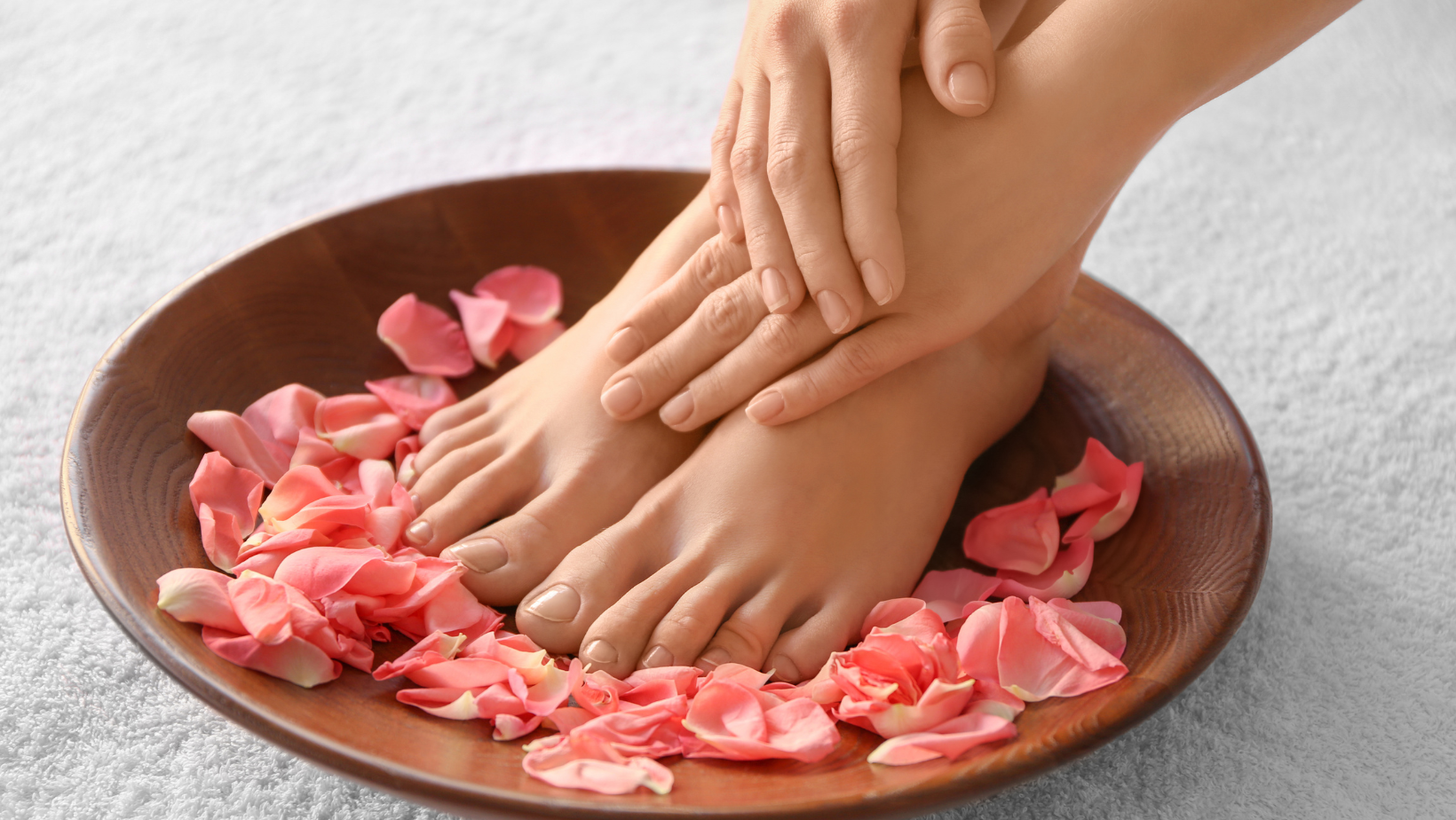 Piedi e mani di donna immersi in una ciotola di legno con petali di rosa rosa, su asciugamano bianco