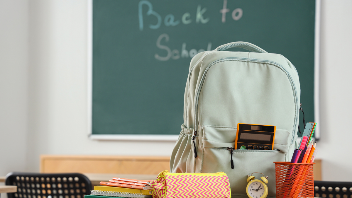 Zaino, astuccio e materiale scolastico su un banco davanti alla lavagna, pronti per il rientro