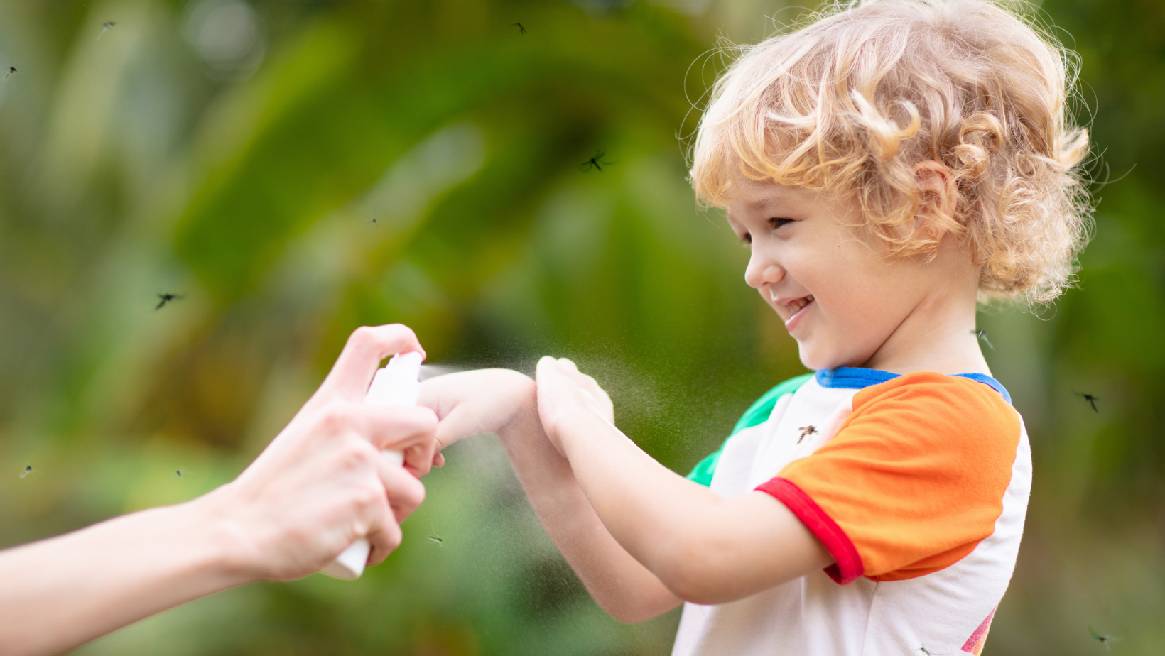 Bambino sorridente all’aperto mentre viene spruzzato con un repellente contro le zanzare.